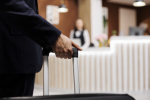 Person in a dark suit pushing a luggage trolley in a hotel lobby with a receptionist in the blurred background